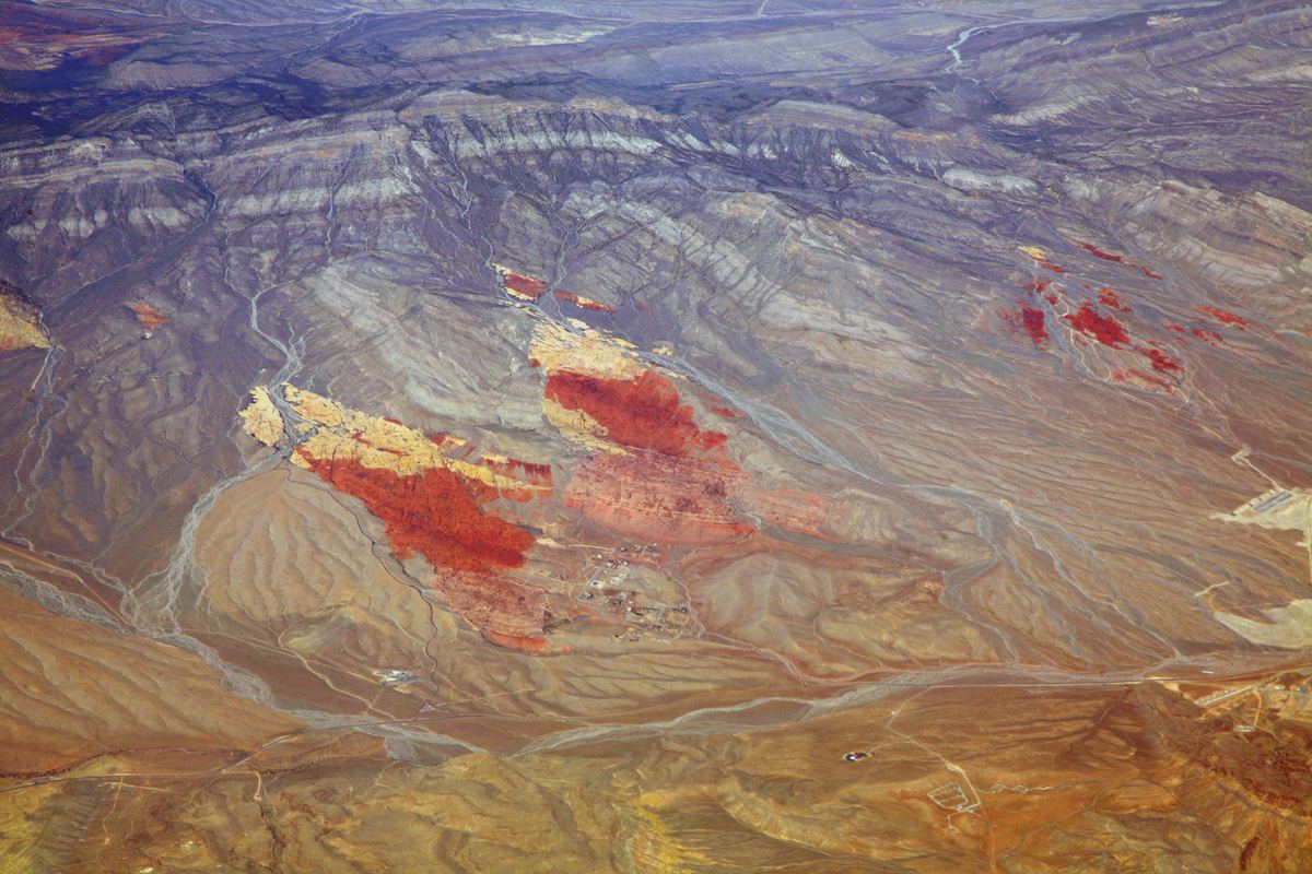 Aerial view of a rugged desert landscape with red and orange rock patches, pale ridges and winding dry stream channels.