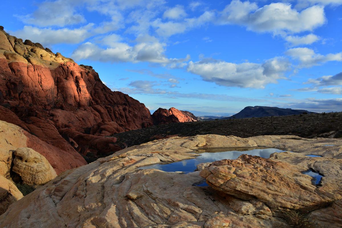 Red and cream sandstone rocks with shallow pools of water in the foreground, dark hills in the distance and a blue sky filled with clouds.