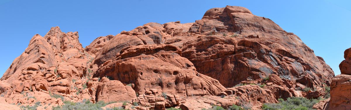 Wide panorama of rugged red sandstone cliffs and rounded rock formations with sparse green shrubs below and a clear blue sky above.