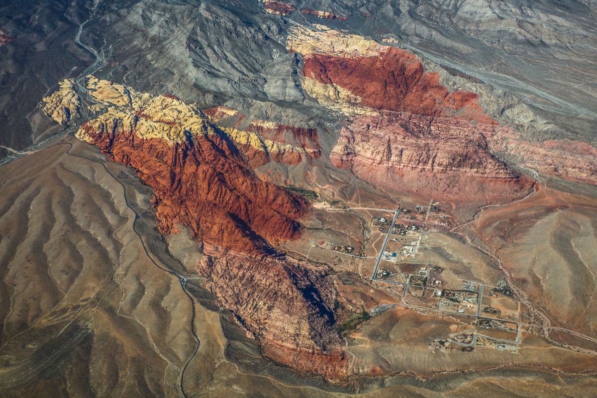 Aerial view of red and tan rock ridges and brown desert slopes, with a small group of buildings and roads on the valley floor.