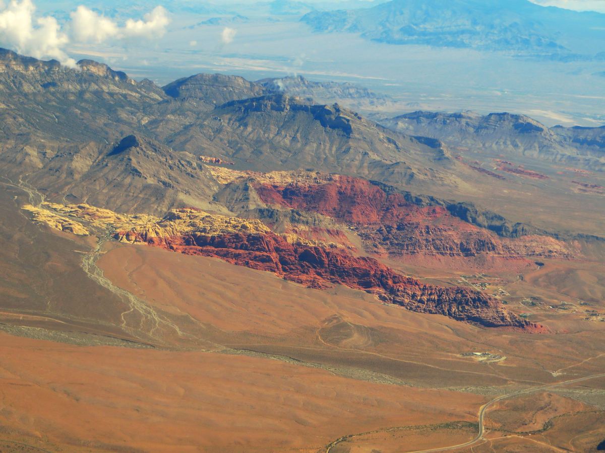 Aerial view of red and tan rock ridges at the edge of a dry desert plain, with rugged mountains and a hazy valley behind.